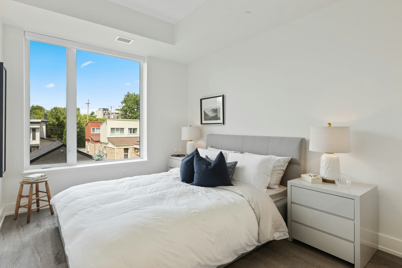 A neatly made bed with white linens and navy pillows in a bright bedroom with a large window